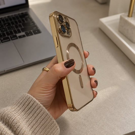 A hand holding a gold-framed smartphone with a transparent back and a circular MagSafe ring, shown in a home office setting with a laptop in the background.