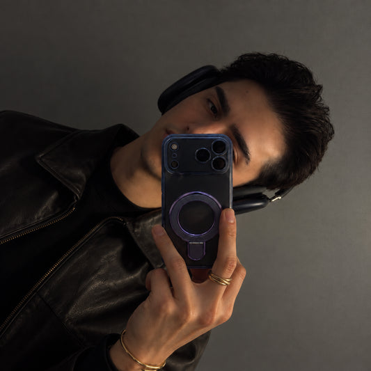 A man wearing a black leather jacket and headphones takes a mirror selfie holding a dark blue MagSafe-compatible smartphone case against a dark gray background.