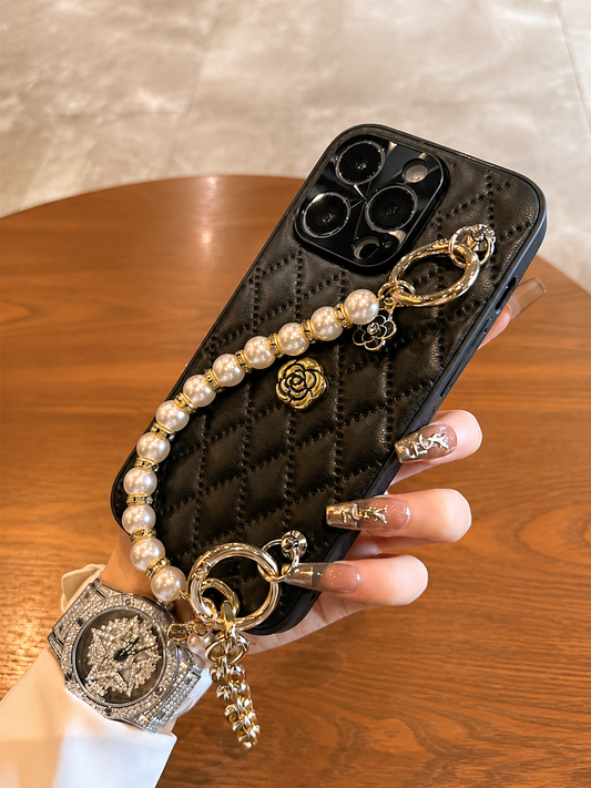 Hand holds the black quilted case above a wooden table. Its gold-colored flower emblem and cream bead strap contrast with the glossy diamond pattern.