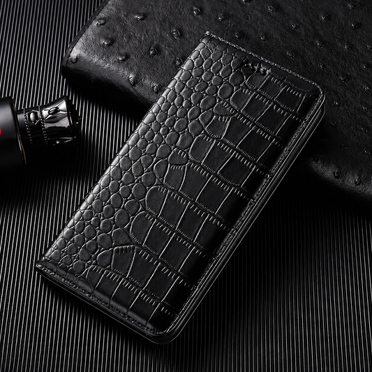 A closed black flip wallet lies diagonally on a dark ribbed surface beside a textured black accessory and small bottle. Its glossy crocodile-like pattern has rounded scales along the fold and larger rectangular scales across the cover. Matching stitches outline the rounded edges.