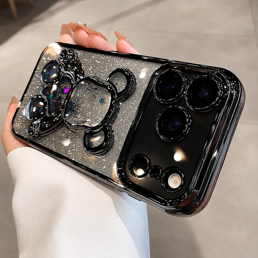 A hand holds a black-trimmed silver-glitter case horizontally indoors, showing a broad three-camera panel and clear bear-shaped decoration filled with multicolored glitter.