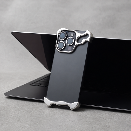 A dark phone with silver sculpted camera and bottom-corner protectors leans against an open black laptop on a gray desk.