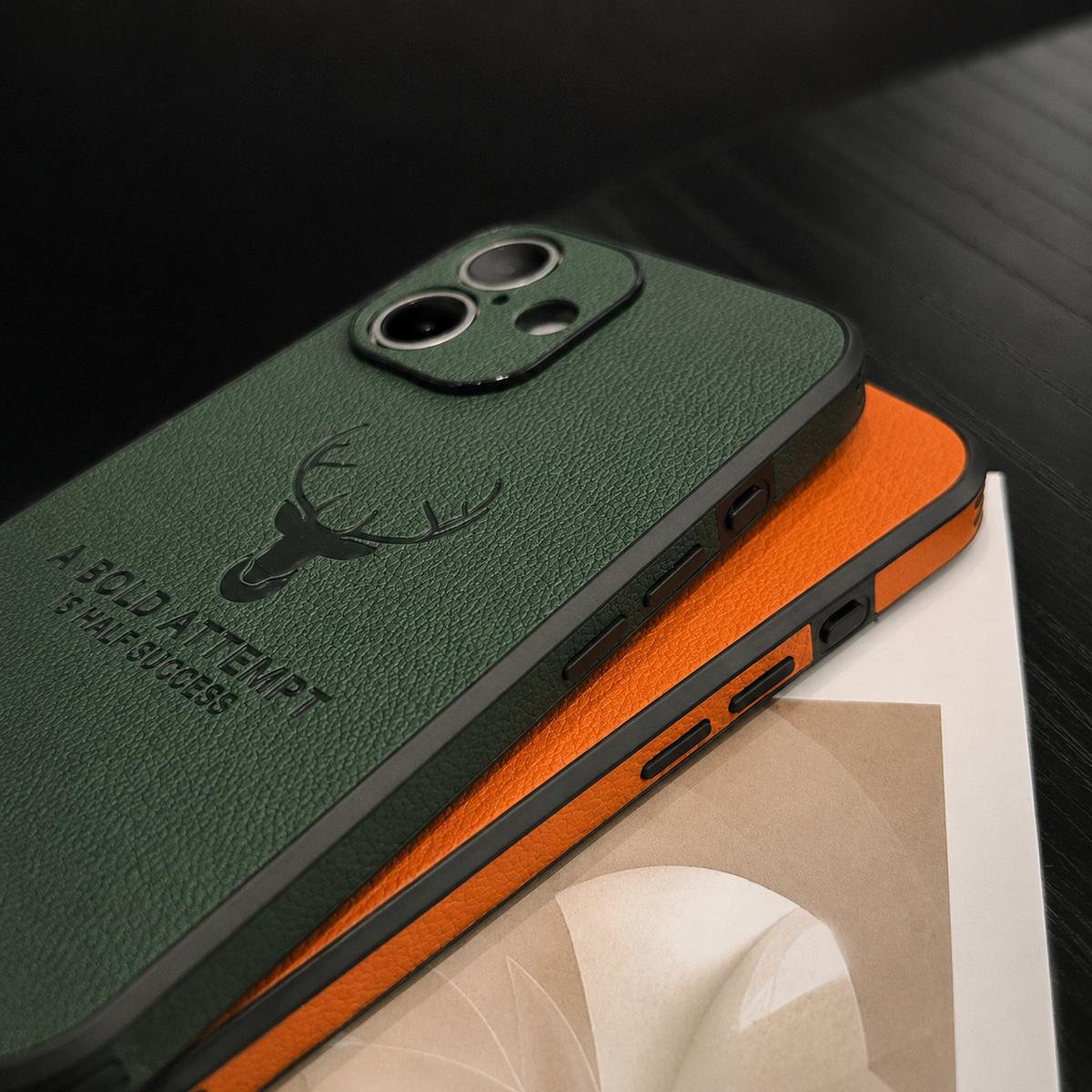 A green case is stacked on an orange case above an open magazine, showing textured backs, deer emblems and side-button openings.