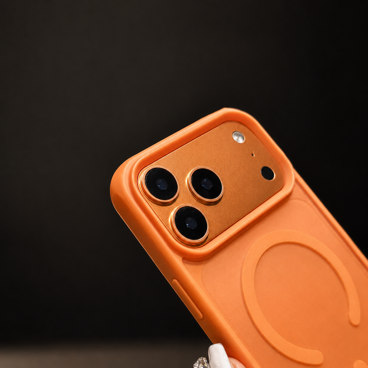 A hand holds an orange case diagonally against black, showing its camera opening and circular magnetic design.