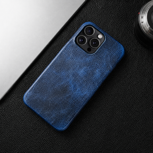 A blue mottled leather-look case lies on a black textured desk beside a laptop and camera lens, showing its small square camera opening.