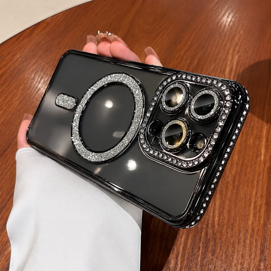 A hand in a white sleeve holds a glossy black phone horizontally above a warm brown wooden tabletop. A sparkling circular ring and short bar decorate the back, while rows of small stones surround the camera panel and phone edges. Three camera lenses sit at the right, reflecting light. Pale manicured fingertips and a gold-colored ring show above the phone.