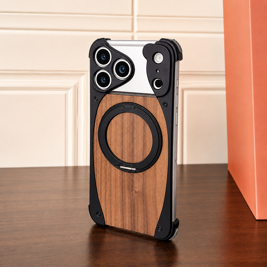 A black metal-looking phone frame with a brown wood-grain back stands on a dark wooden table beside an orange box. A matching circular folding ring lies flat over the wood panel. Screws secure reinforced rounded corners, and separate raised openings surround the cameras and sensors.