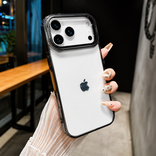 Hand holds a clear black-edged case in an indoor setting with a wooden counter behind it. A broad raised rectangular frame surrounds the phone's three cameras.