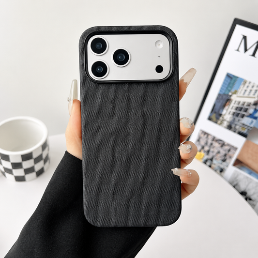 A hand in a black sleeve holds a black fabric-textured phone case upright above a white tabletop. A pale rectangular camera panel spans the top, showing three black lenses and smaller openings. A black-and-white checkered mug sits behind the hand at left, and a partly cropped magazine lies at right. The long fingernails are pale.