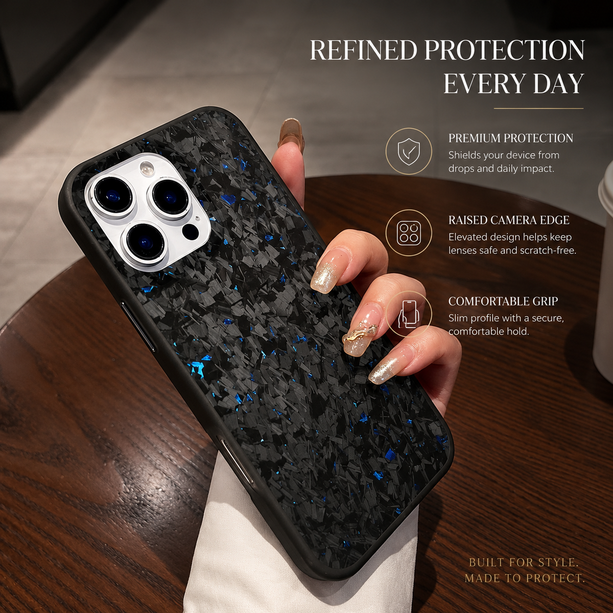 A hand with long pale decorated nails holds a blue-flecked black phone diagonally above a wooden table. A pale rectangular camera panel with three lenses sits at the upper end, surrounded by the mottled case. A white sleeve covers the wrist. 'Refined Protection Every Day' appears above three feature labels at right.
