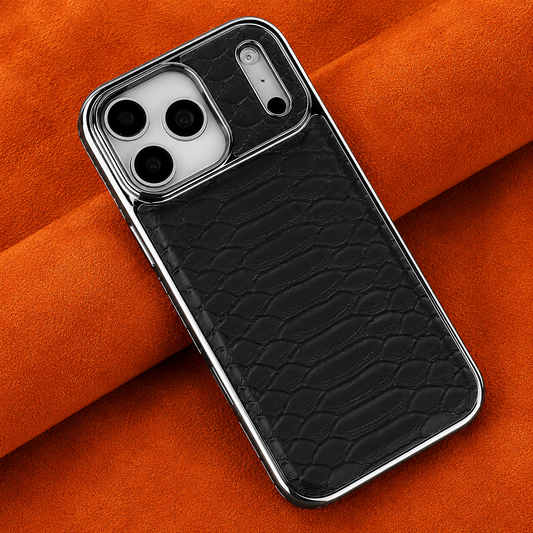 A black snakeskin-patterned phone lies diagonally across folded orange fabric. Its raised scale texture is framed by a polished silver-colored border. Three camera lenses sit in a pale panel at the upper end, beside a narrow silver-colored strip with small round details. Soft folds cast shadows beneath the phone.