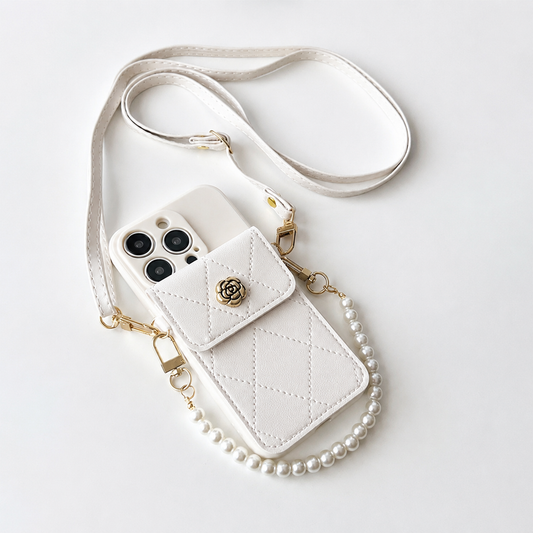 An ivory quilted phone wallet lies diagonally on a pale surface beside its long matching shoulder strap. Diamond stitching covers the rear pocket, which closes with a flap and gold rose-shaped ornament. A short strand of pearl-like white beads attaches to gold clips at the sides, forming a small carrying handle.