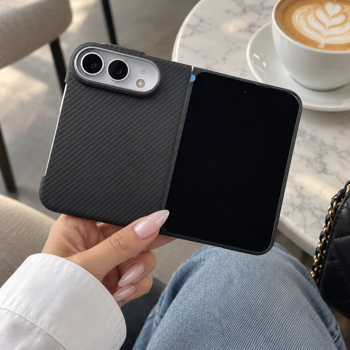 A person holds a black carbon-fiber-textured foldable phone case with a black smartphone inside, photographed in a cafe setting with a latte and table visible in the background.