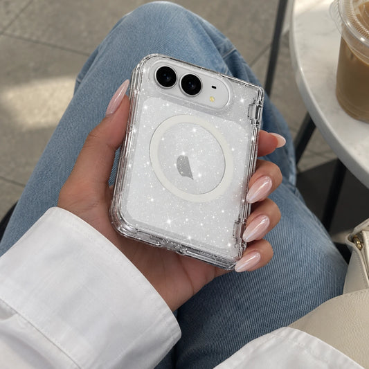 A hand holding a clear glitter phone case with a MagSafe ring, shown on a phone. The person is wearing blue jeans and a white shirt, with a coffee cup on a table in the background.