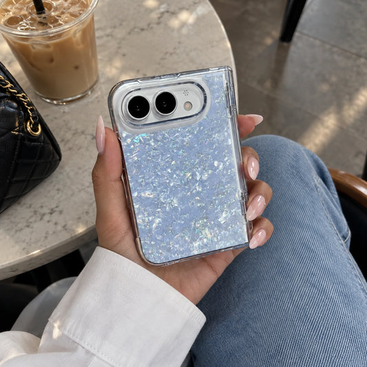 A person holding a clear phone case with iridescent glitter in a light blue shade, seated at a cafe table with an iced coffee and a black quilted handbag nearby.