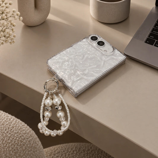 A silver metallic textured phone case with a pearl-bead strap ring, resting on a beige desk next to a laptop, mug, and decorative vase.