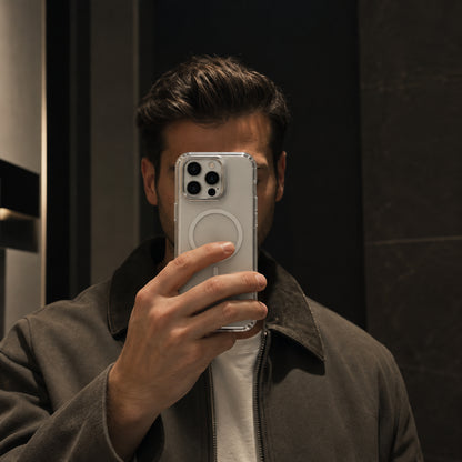 A man takes a mirror selfie showing a clear protective phone case on a silver smartphone, wearing a brown jacket over a white t-shirt against a dark background.