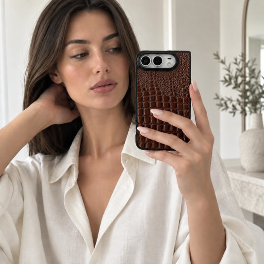 A woman takes a mirror selfie showing off a dark brown crocodile-embossed phone case. She is wearing a white linen shirt, and a neutral-toned interior room with a plant is visible in the background.