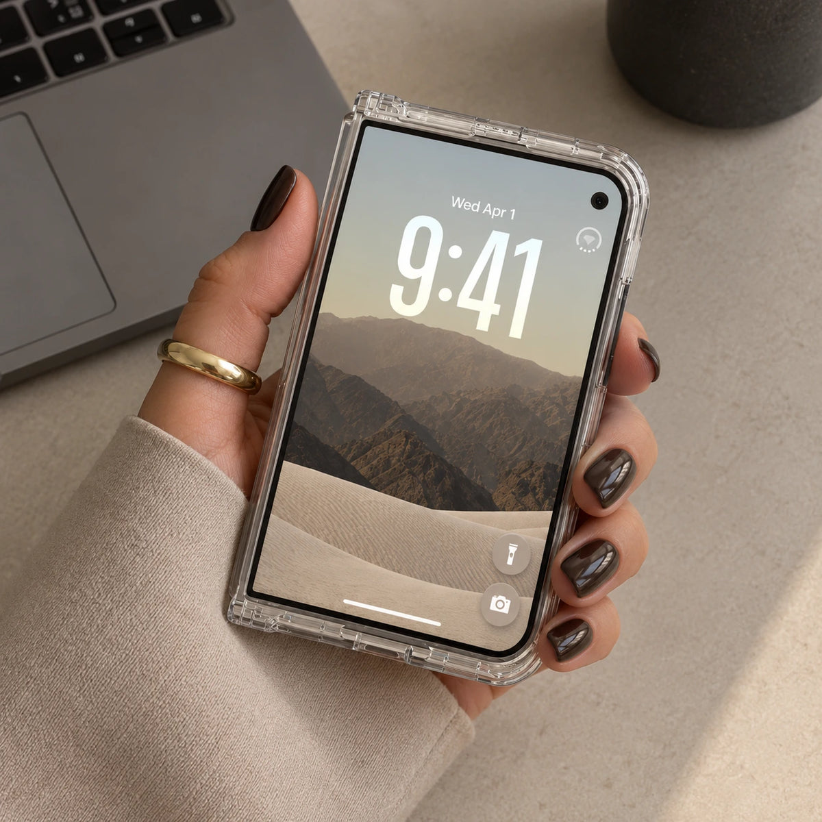 A hand with dark glittery nail polish holds a smartphone in a clear, transparent bumper case. The phone displays a mountain landscape wallpaper with the time 9:41.