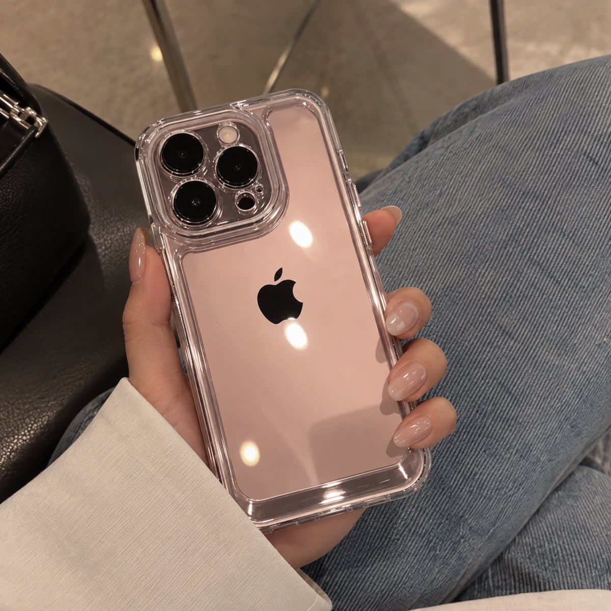 A clear protective phone case with a black camera border, shown on an iPhone held in a hand. The person is wearing blue jeans and a white long-sleeve top.