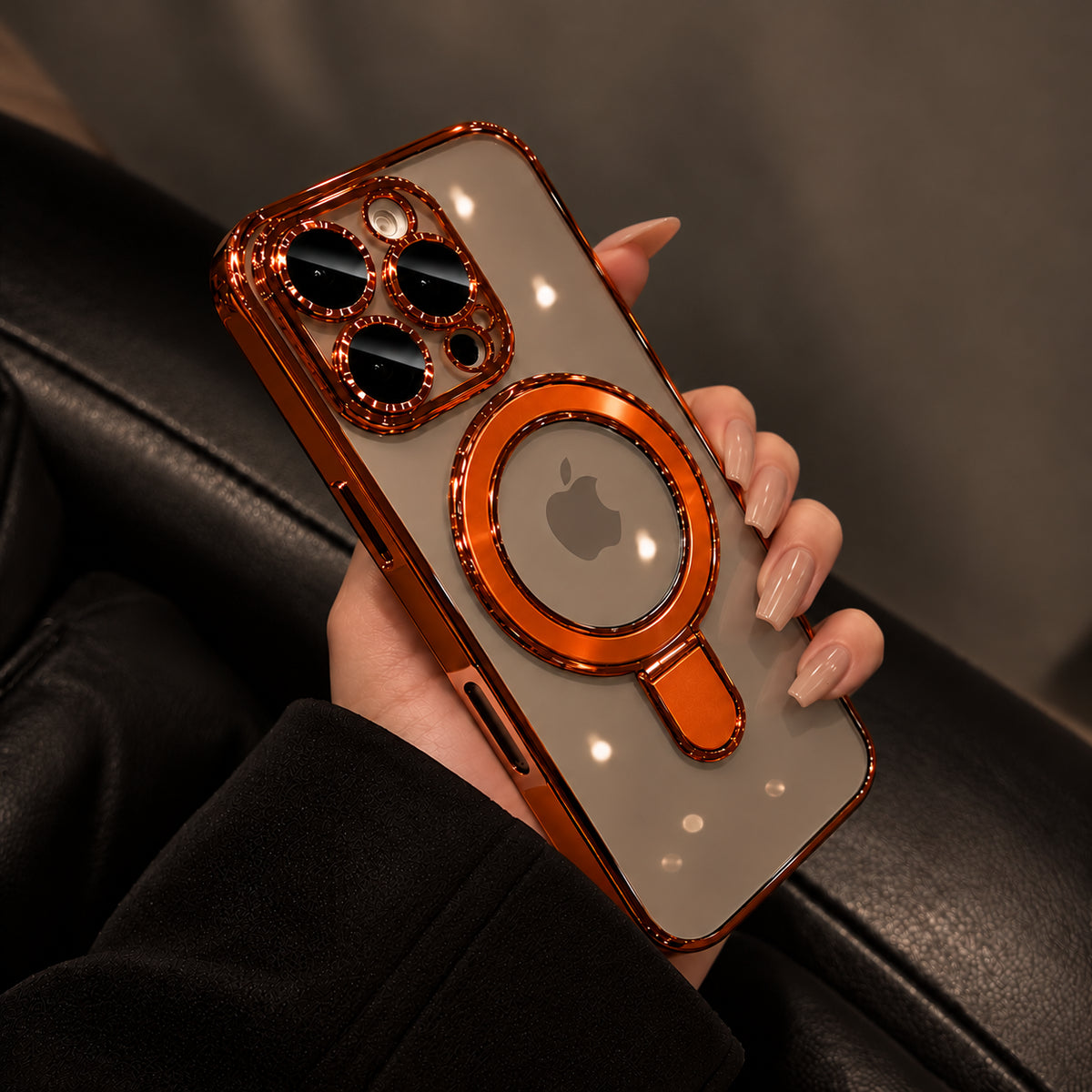 A hand with manicured nails holds a smartphone in a black leather chair, showing an ornate phone case. The case has a rose gold metallic frame, a transparent back panel, a built-in magnetic ring, and a magnifying glass design around the camera lenses.