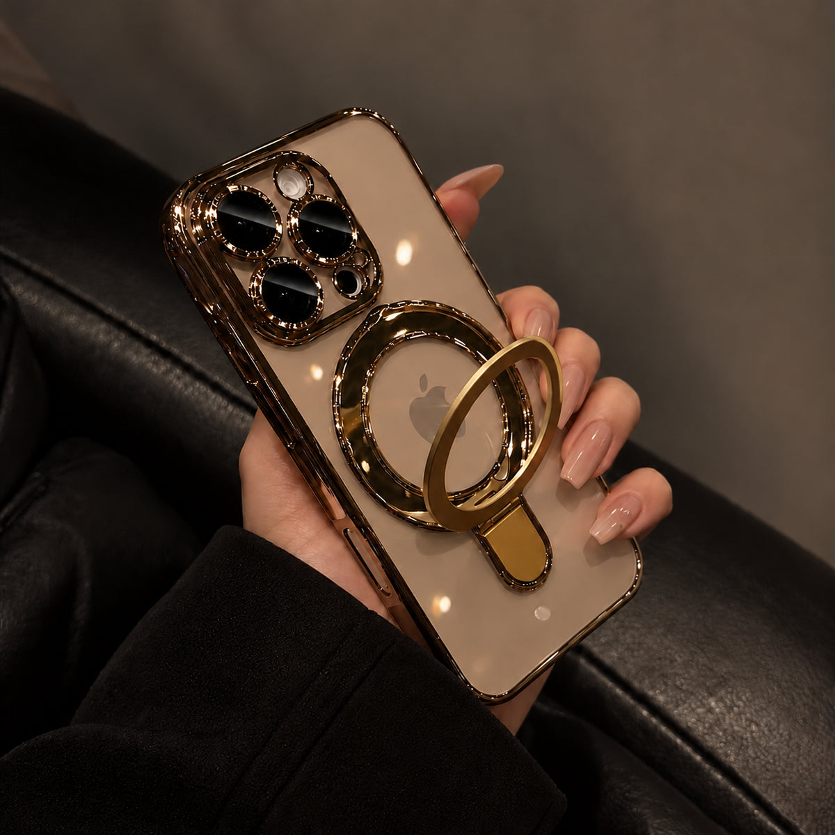 A hand with manicured nails holds a smartphone in a gold-toned case with a circular ring stand. The phone has a frosted beige back panel and a metallic gold camera surround, photographed against a dark leather seat.