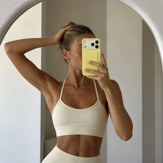 A woman takes a mirror selfie wearing a cream-colored ribbed sports bra and matching high-waisted leggings, holding a yellow smartphone case.