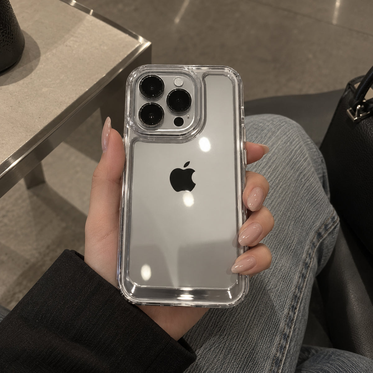 A clear, transparent protective phone case fitted on an iPhone, held by a hand wearing a black long-sleeve top and light blue jeans in a casual indoor setting.