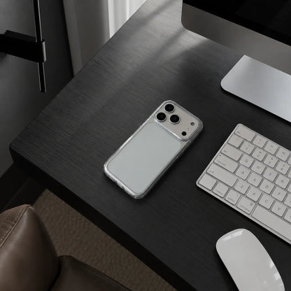 A clear smartphone case with a silver metallic back and camera cutout, shown on a dark wood desk next to a keyboard, mouse, and computer monitor.