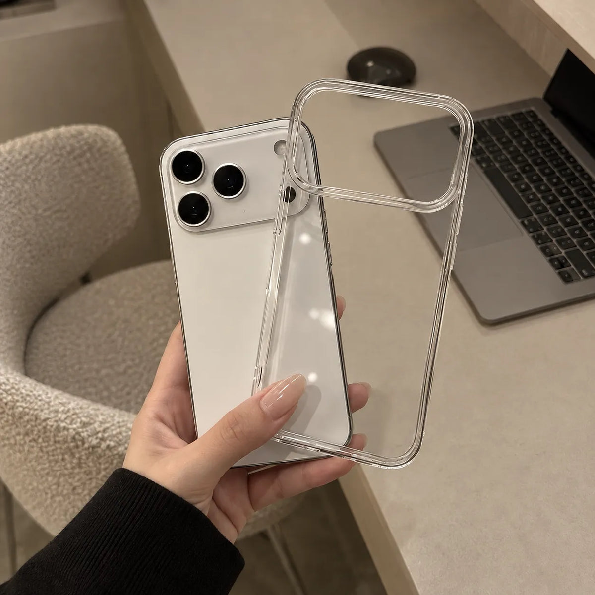 A hand holds a clear, transparent phone case over a white smartphone, with a desk, laptop, and chair visible in the background.