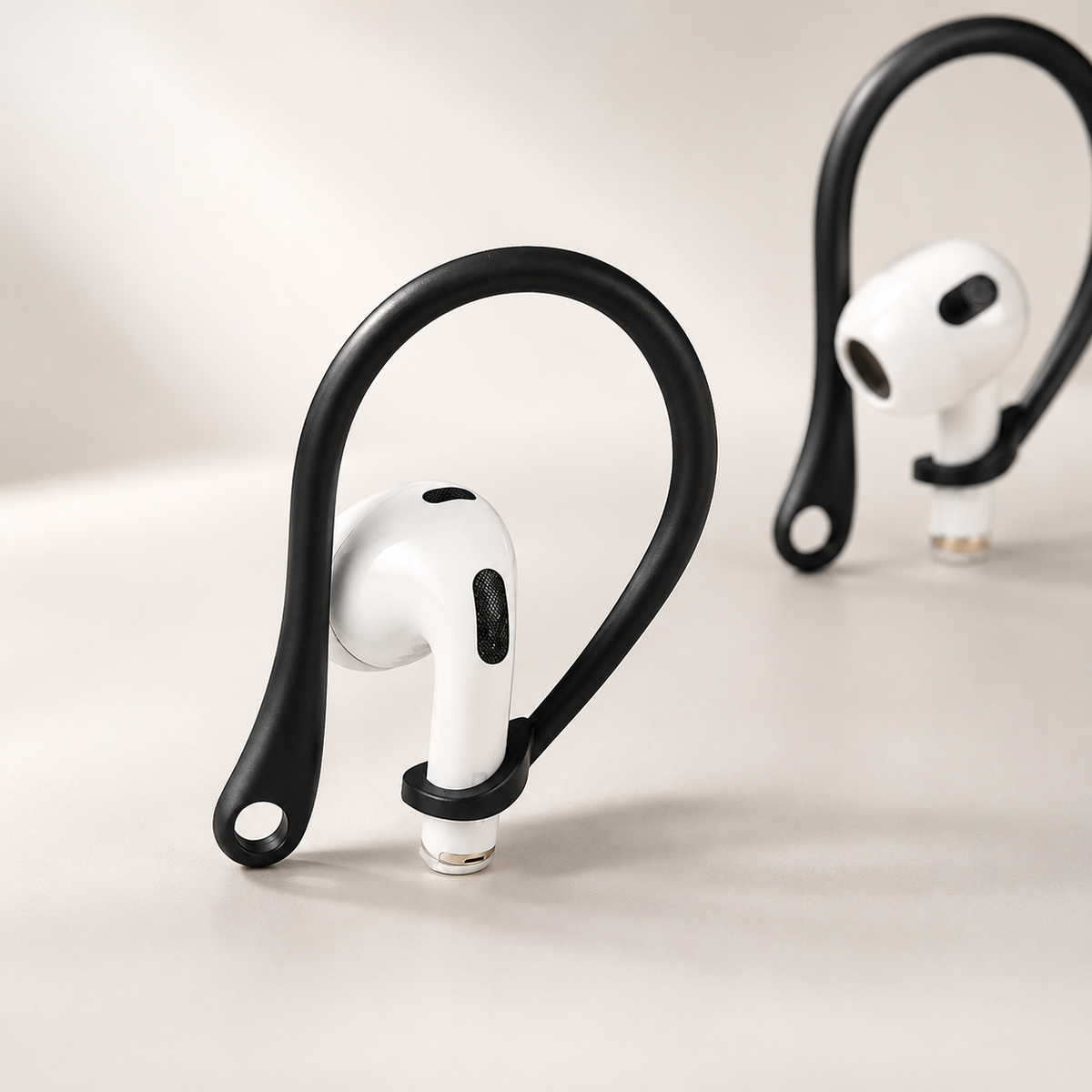 Two black curved ear hooks fitted to white stemmed earbuds stand on a beige surface. Each loop arches above the earbud and ends in a small round opening; the nearer earbud faces forward and the other sits farther back at an angle.