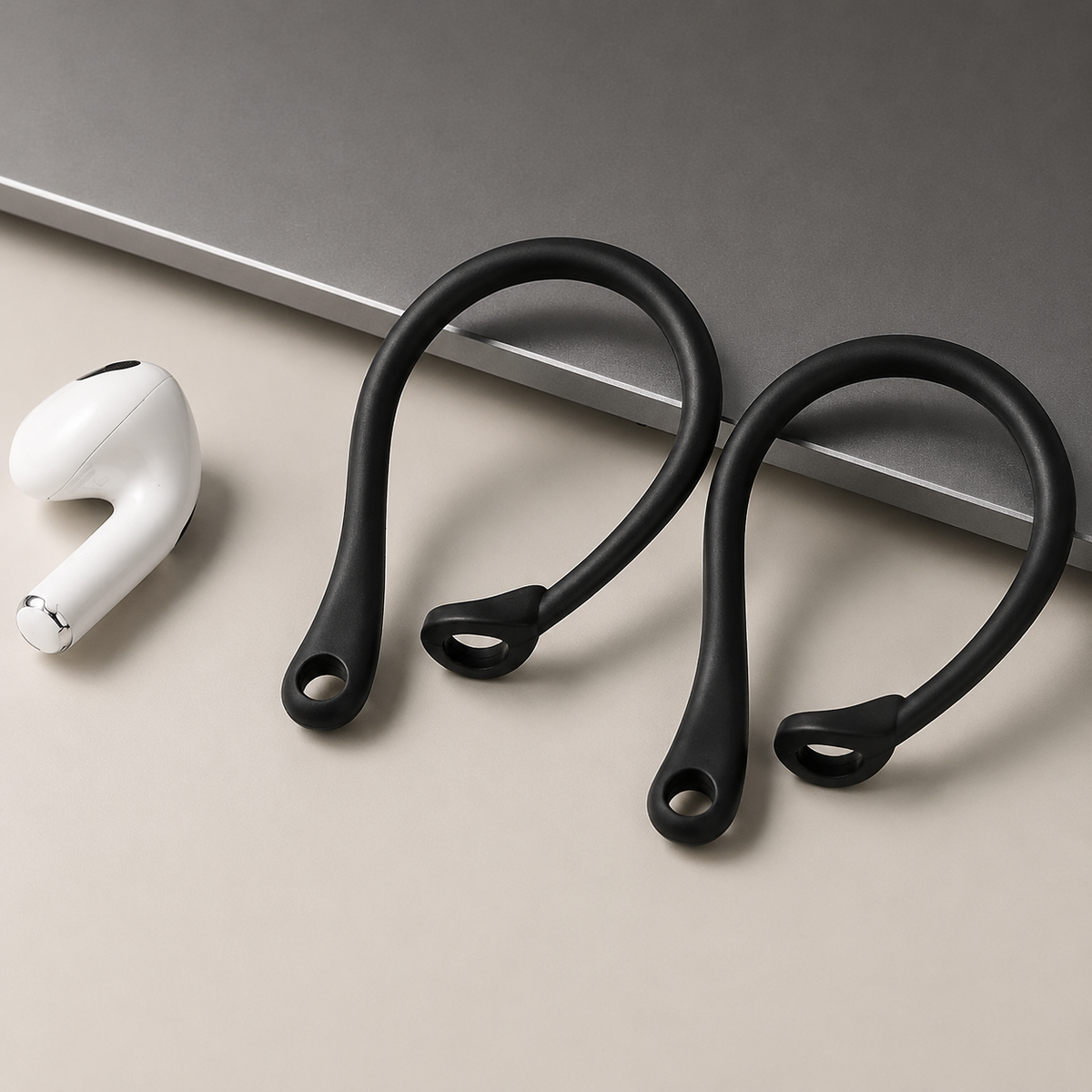 Two empty black ear hooks lie on a beige surface beside a loose white earbud and the corner of a gray laptop. Their long curved loops and small circular collars for gripping the earbud stems are clearly visible.