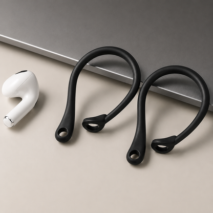 Two empty black ear hooks lie on a beige surface beside a loose white earbud and the corner of a gray laptop. Their long curved loops and small circular collars for gripping the earbud stems are clearly visible.
