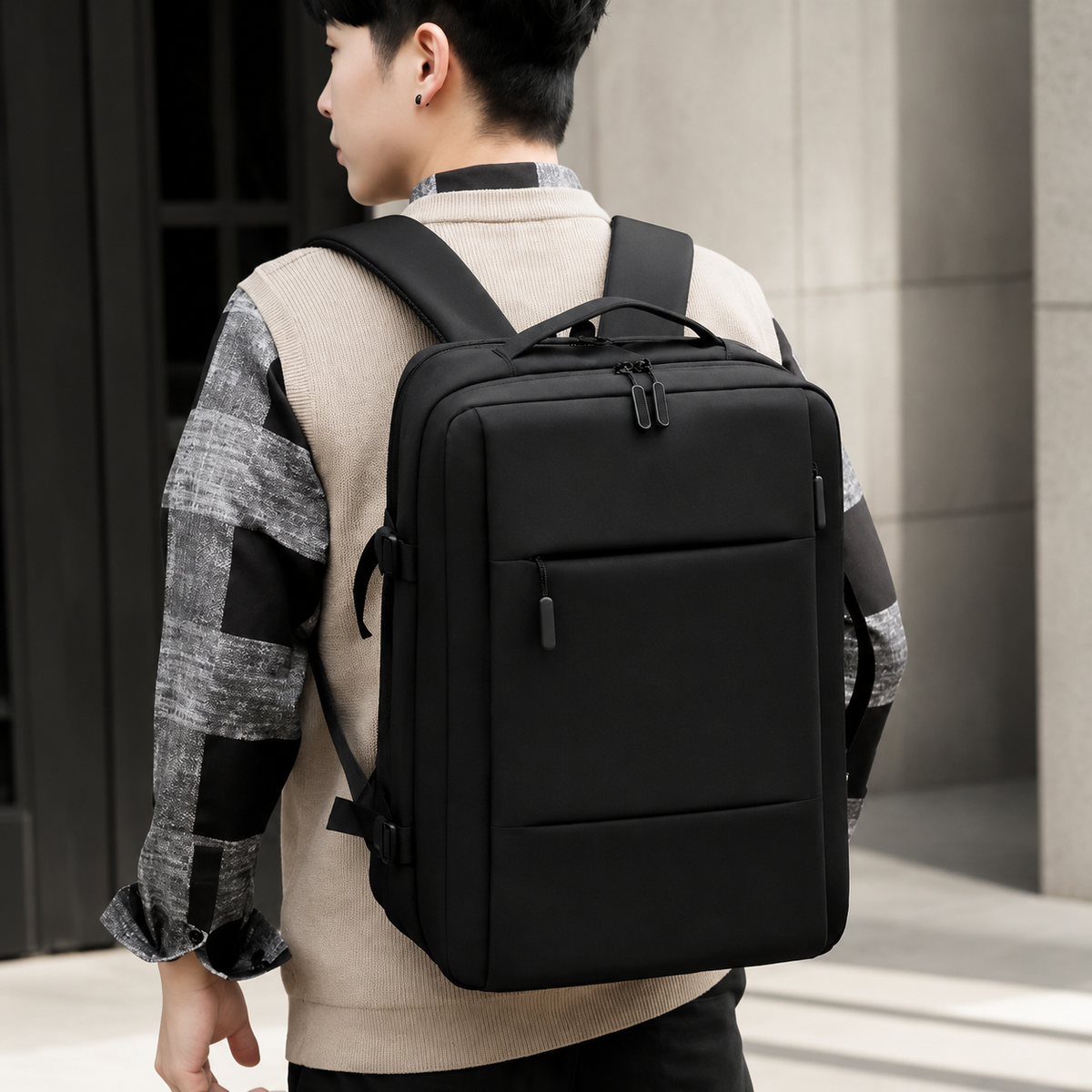 A man seen from behind wears a black rectangular travel backpack over a gray patterned shirt and beige vest. The bag has a top handle, front zip pockets, padded shoulder straps and small side compression buckles. Pale building columns form the background.