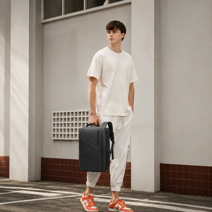 A man in a loose white T-shirt, pale trousers and orange sneakers stands outside a building, carrying a black rectangular travel bag by its top handle. The bag hangs beside his leg, showing its slim shape and rounded corners.
