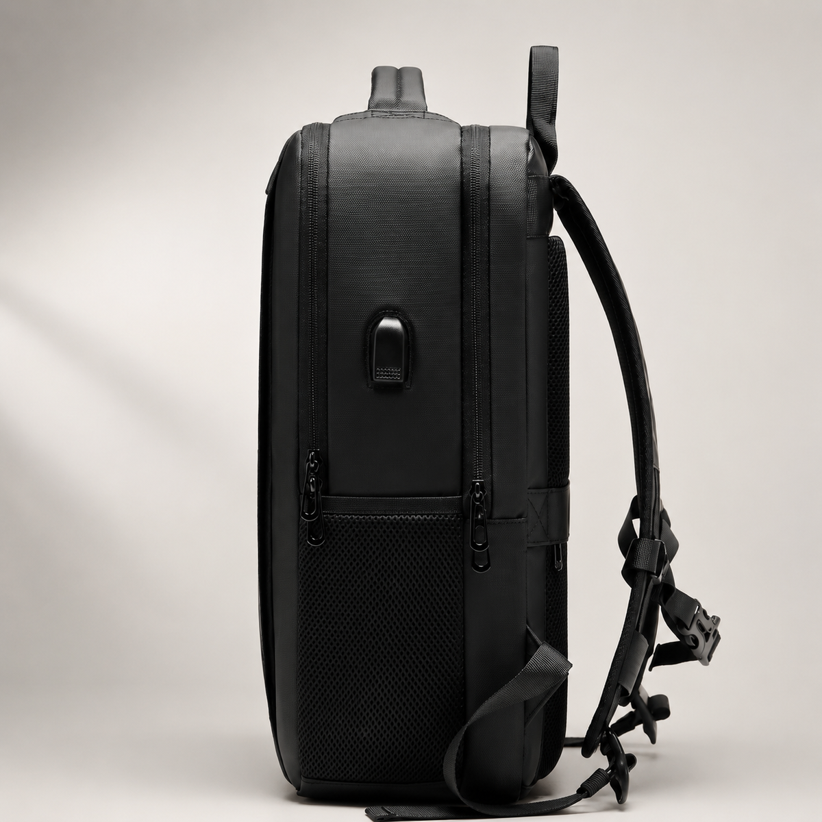 Side view of a black travel backpack shows several zipper tracks, a small USB port, a padded side carry handle and a curved shoulder strap extending behind it. The upright bag has a thick rectangular profile against a pale background.