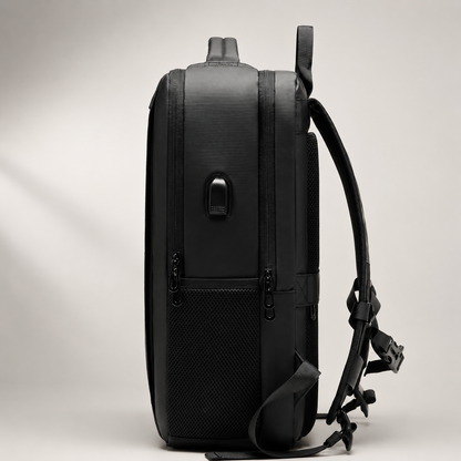 Side view of a black travel backpack shows several zipper tracks, a small USB port, a padded side carry handle and a curved shoulder strap extending behind it. The upright bag has a thick rectangular profile against a pale background.