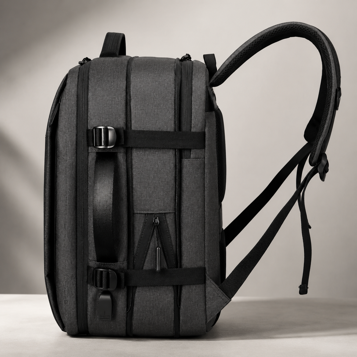 A charcoal backpack's side shows stacked zippered compartments, compression straps, side handle and a curved padded shoulder strap.