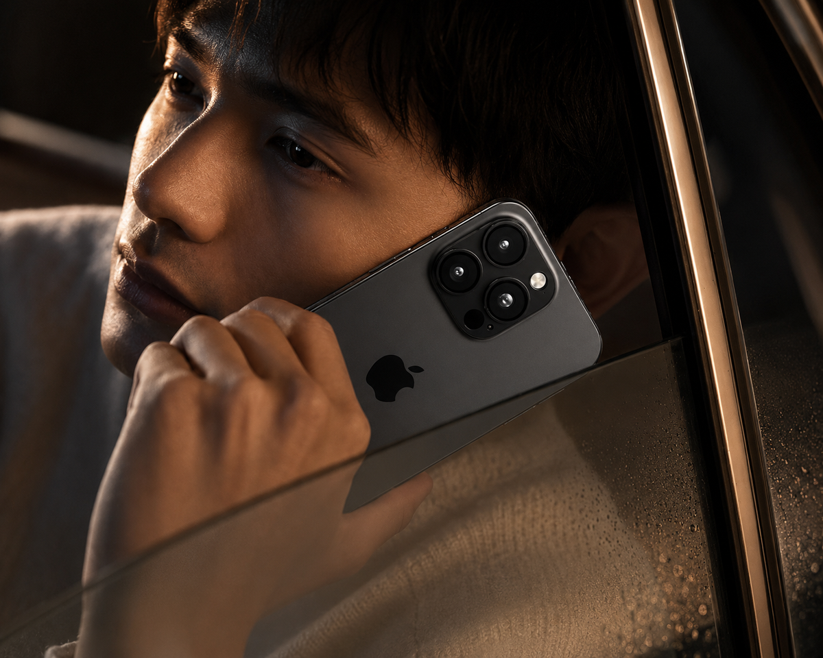 A man holds a dark phone against his cheek while sitting beside a window. The phone's rear camera cluster and fitted lens rings are visible.