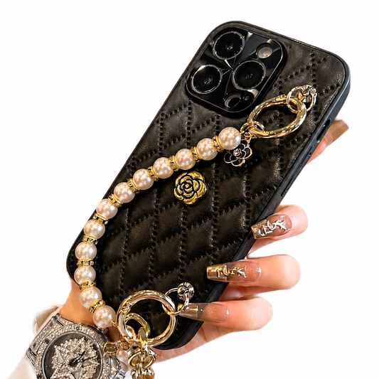 Hand holds a black diamond-quilted case with a small gold-colored flower emblem. A cream pearl-like bead strap with gold-colored links and dangling charms arcs across the back.