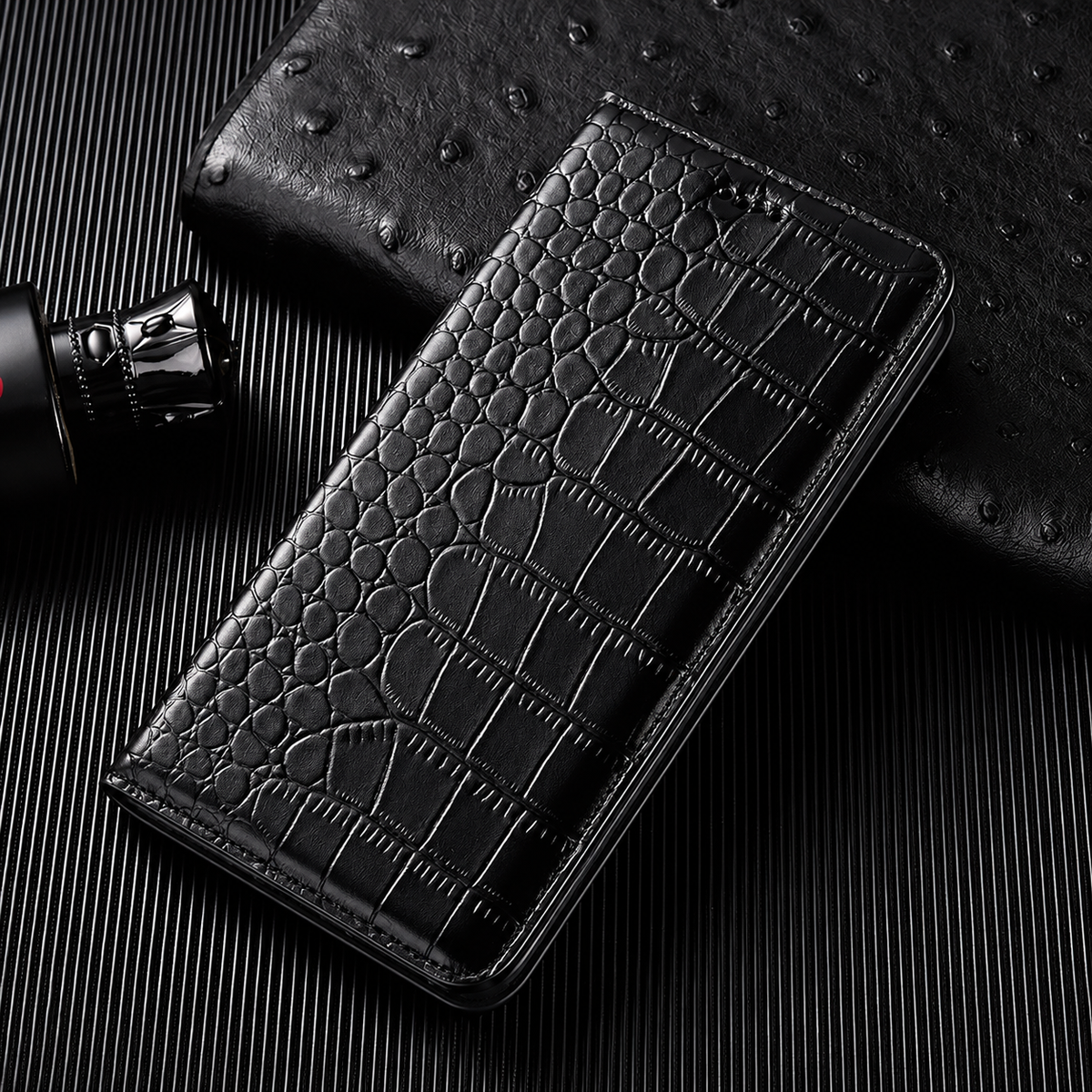 A closed black flip wallet lies diagonally on a dark ribbed surface beside a textured black accessory and small bottle. Its glossy crocodile-like pattern has rounded scales along the fold and larger rectangular scales across the cover. Matching stitches outline the rounded edges.
