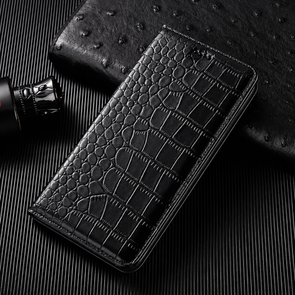 A closed black flip wallet lies diagonally on a dark ribbed surface beside a textured black accessory and small bottle. Its glossy crocodile-like pattern has rounded scales along the fold and larger rectangular scales across the cover. Matching stitches outline the rounded edges.