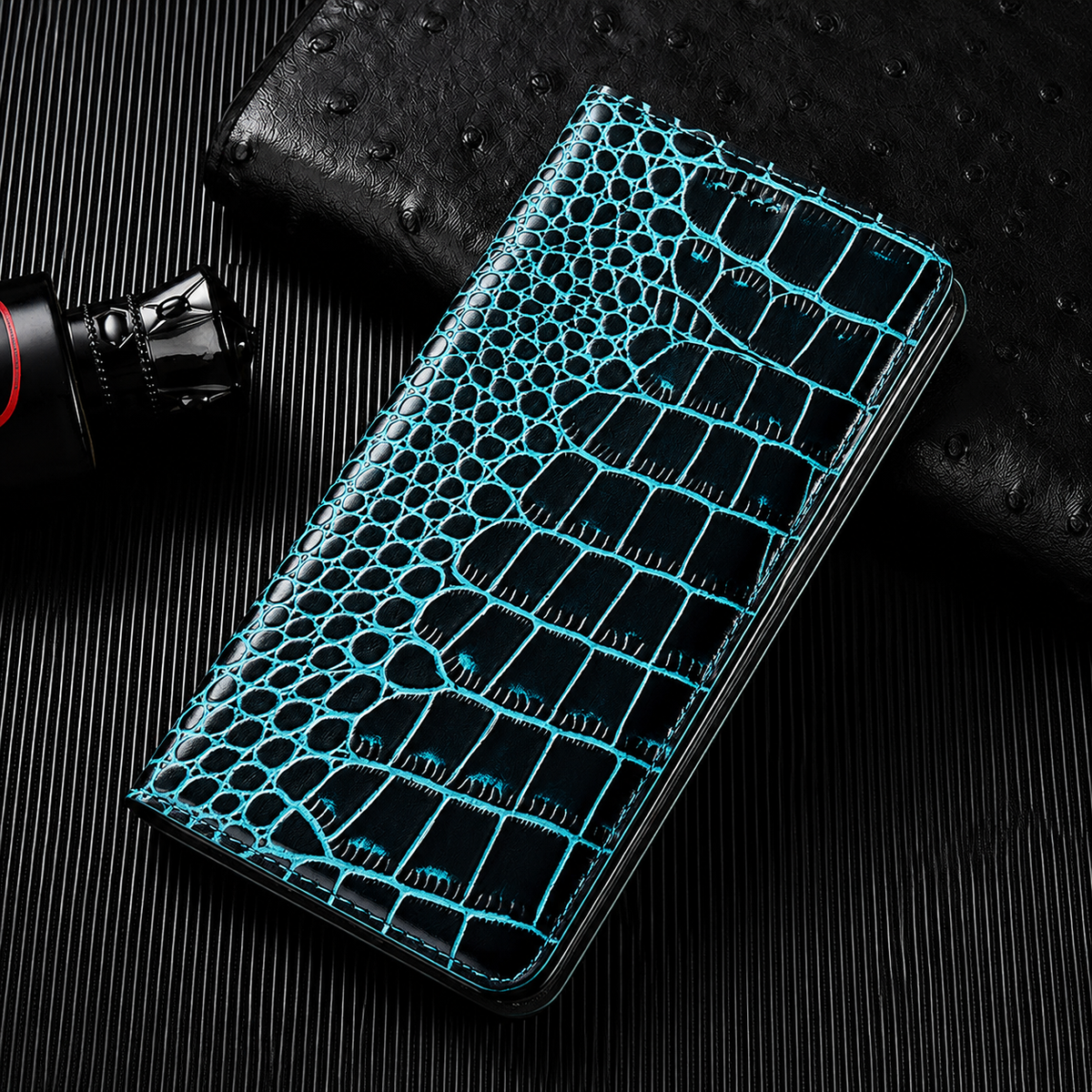 A closed black flip wallet with turquoise scale outlines lies diagonally on a dark ribbed surface beside a textured black accessory and small bottle. Its glossy crocodile-like pattern has rounded scales along the fold and larger rectangular scales across the cover. Matching stitches outline the rounded edges.