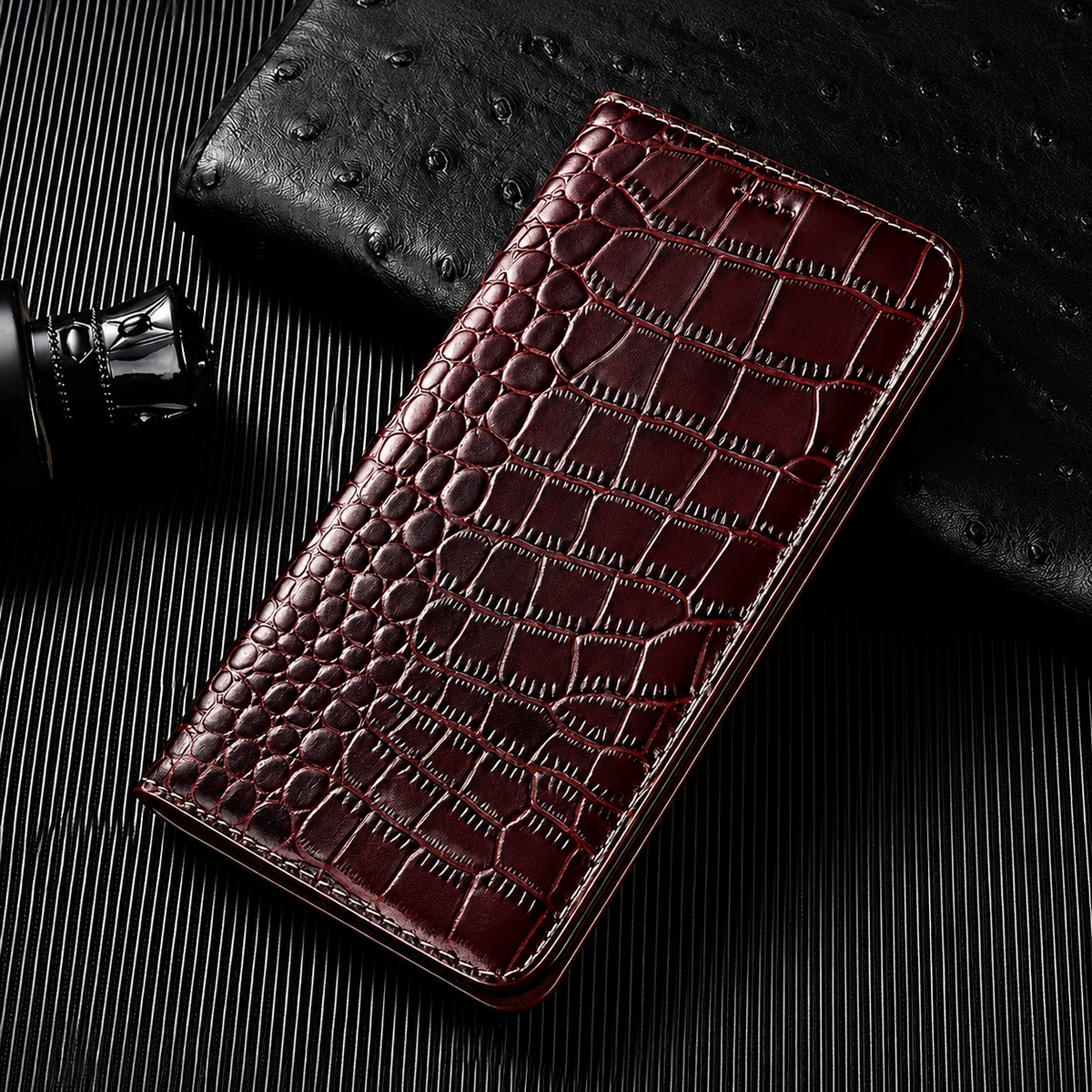 A closed burgundy flip wallet lies diagonally on a dark ribbed surface beside a textured black accessory and small bottle. Its glossy crocodile-like pattern has rounded scales along the fold and larger rectangular scales across the cover. Matching stitches outline the rounded edges.