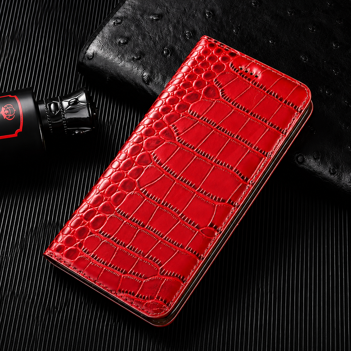 A closed bright red flip wallet lies diagonally on a dark ribbed surface beside a textured black accessory and small bottle. Its glossy crocodile-like pattern has rounded scales along the fold and larger rectangular scales across the cover. Matching stitches outline the rounded edges.
