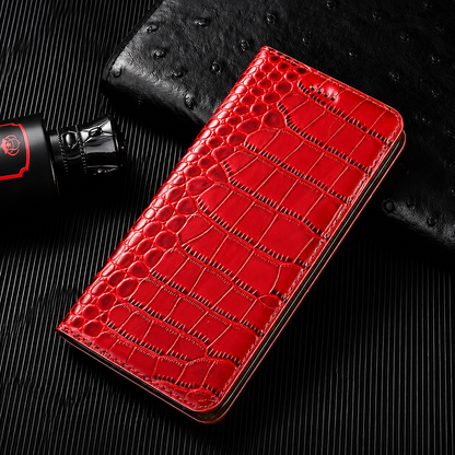 A closed bright red flip wallet lies diagonally on a dark ribbed surface beside a textured black accessory and small bottle. Its glossy crocodile-like pattern has rounded scales along the fold and larger rectangular scales across the cover. Matching stitches outline the rounded edges.