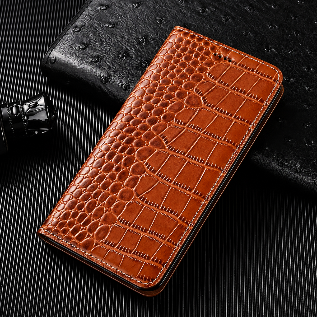 A closed tan flip wallet lies diagonally on a dark ribbed surface beside a textured black accessory and small bottle. Its glossy crocodile-like pattern has rounded scales along the fold and larger rectangular scales across the cover. Matching stitches outline the rounded edges.