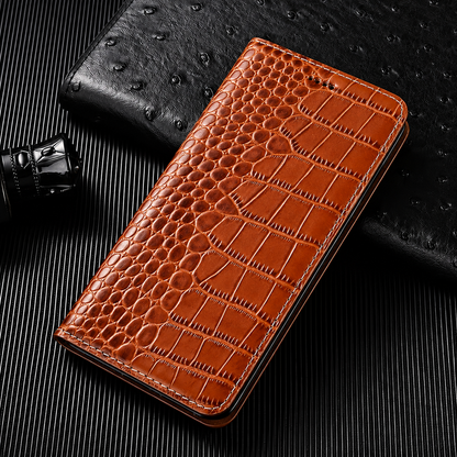 A closed tan flip wallet lies diagonally on a dark ribbed surface beside a textured black accessory and small bottle. Its glossy crocodile-like pattern has rounded scales along the fold and larger rectangular scales across the cover. Matching stitches outline the rounded edges.