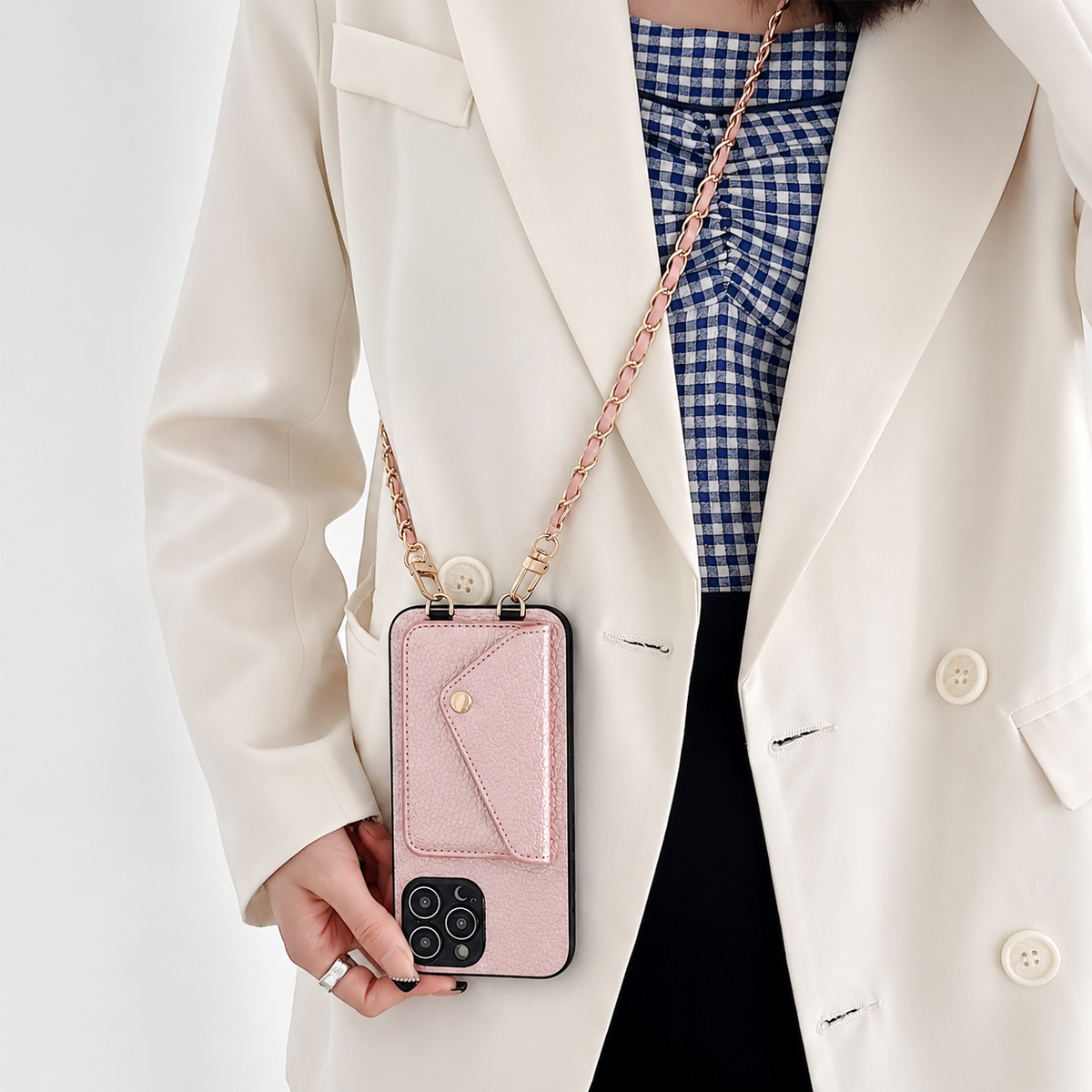 Pink pebbled phone wallet hangs vertically from a gold-colored chain against a woman's cream blazer. Its envelope-shaped rear pocket is closed above the exposed cameras.