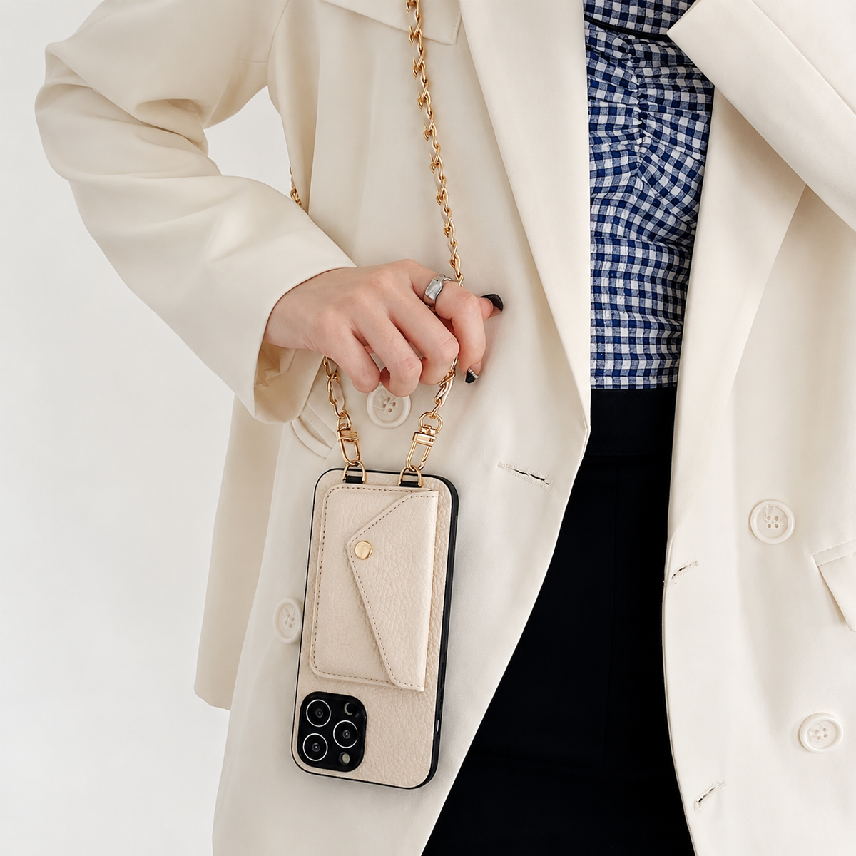 A woman in a cream blazer holds the chain of a cream phone wallet hanging vertically. A gold-colored snap fastens its envelope-shaped rear pocket.